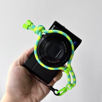 Hand holding a camera with a green and yellow braided strap on a light gray background