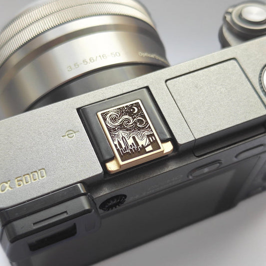 Close-up of a camera body with a textured silver and black finish, featuring a decorative hot shoe cover with a swirling starry sky design and gold trim.
