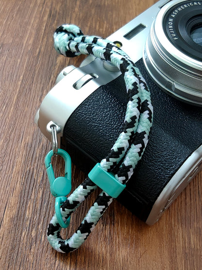 Camera with a braided strap featuring black and white patterns and a turquoise carabiner on a wooden surface.