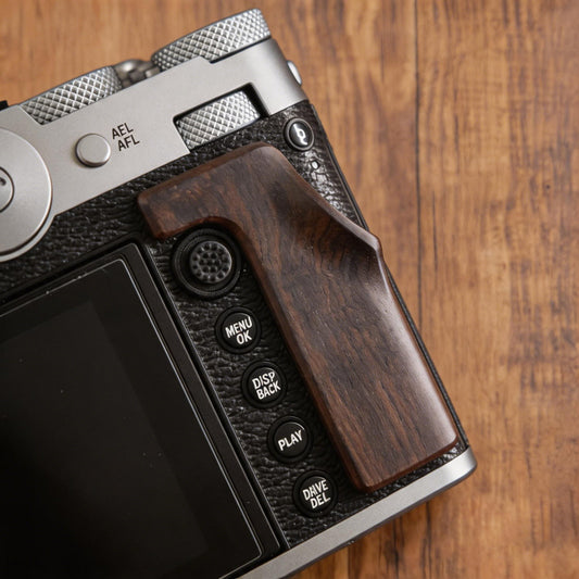 A close-up of a Fuji GFX100RF camera body with a dark brown wooden grip attachment, resting on a wooden surface. 