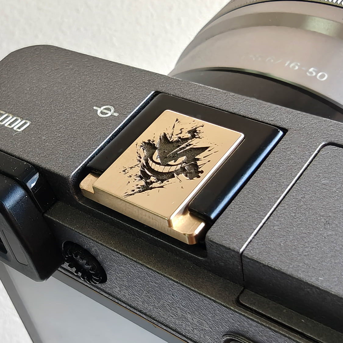 Close-up of a camera with a gold gengar hot shoe cover on a light gray background