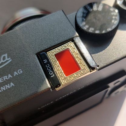 A gold-colored metal camera hot shoe cover placed on a black camera, focusing on the cover with the brand name visible.