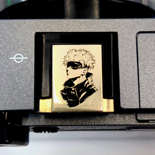 A gold hot shoe cover featuring gojosatoru attached to the front of a camera.