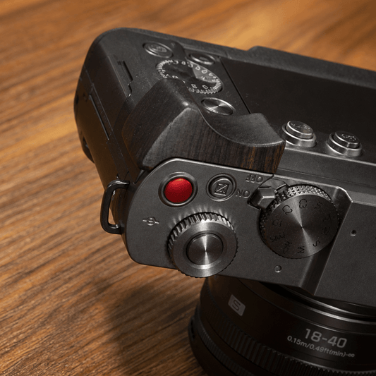 Close-up of a camera body showing a dark wood grip insert, a red record button, silver metal dials, and engraved markings, with part of a lens visible against a white background.